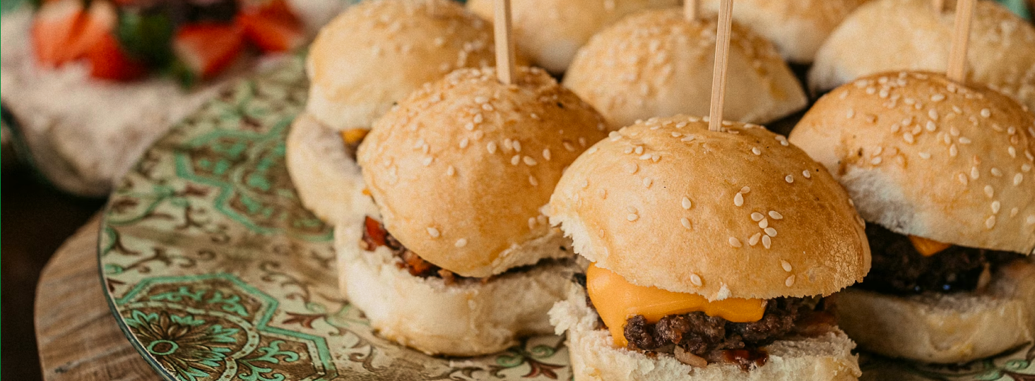 Mini burgery z roztopionym serem w sezamowych bułkach, nadziane na patyczki na ozdobnym talerzu.