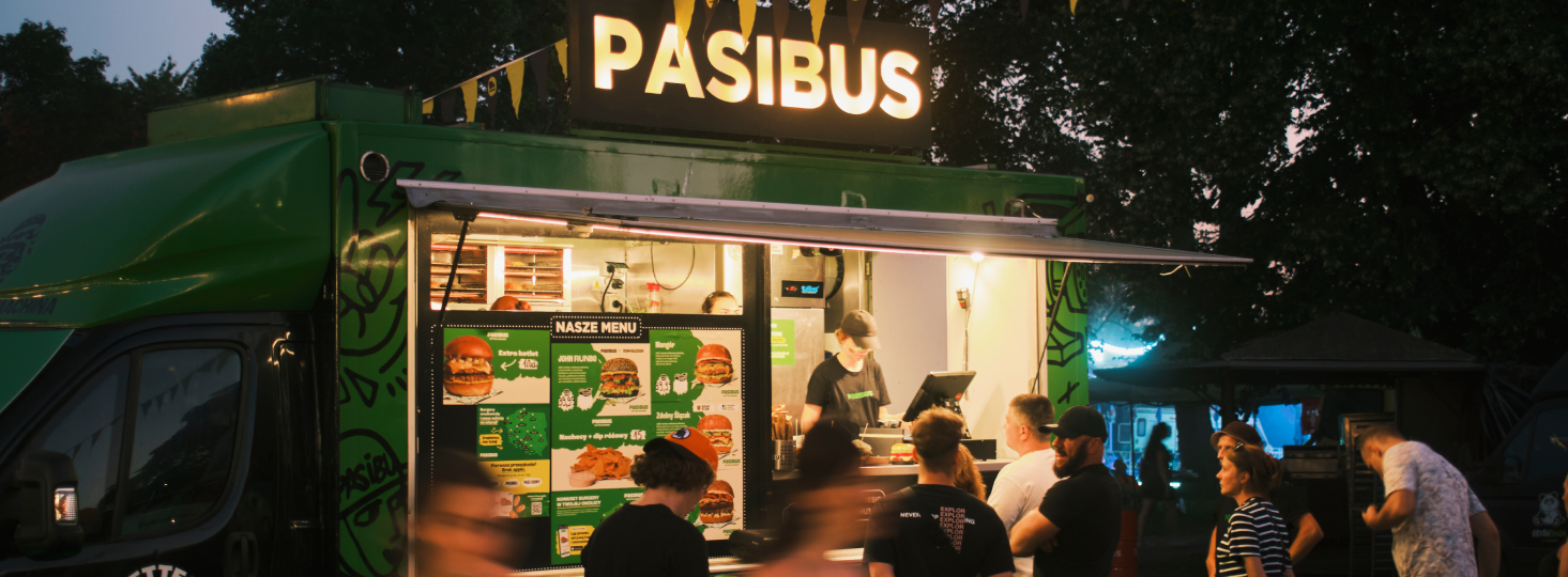 Zielony food truck Pasibus z podświetlanym napisem i klientami zamawiającymi burgery wieczorem.